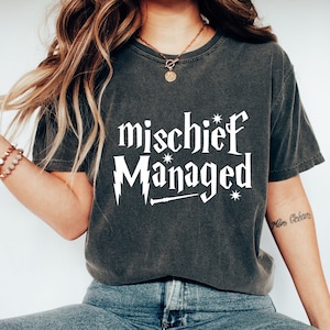 May include: A dark gray t-shirt with the text "mischief Managed" in white lettering. The text is stylized to look like a Harry Potter themed graphic. The shirt is being worn by a person with long brown hair.