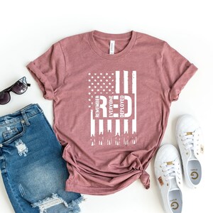 Könnte beinhalten: Ein mauvefarbenes T-Shirt mit dem Design der amerikanischen Flagge und den weißen Worten "REMEMBER EVERYONE DEPLOYED RED". Das Shirt wird mit blauen Jeansshorts, weißen Turnschuhen und einer schwarzen Sonnenbrille kombiniert. Die Artikel sind auf einer weißen Oberfläche angeordnet.