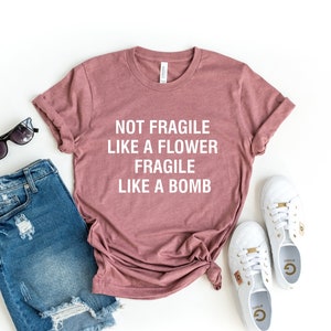 Puede incluir: Una camiseta color malva con el texto "NOT FRAGILE LIKE A FLOWER FRAGILE LIKE A BOMB" en blanco. La camiseta está colocada sobre un fondo blanco con unos pantalones cortos de mezclilla desgastados, gafas de sol y zapatillas blancas.