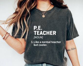 May include: Dark gray t-shirt with the text "P.E. TEACHER (NOUN) 1. Like a normal teacher but cooler." in white. The shirt is worn with blue jeans and white sneakers. A gold necklace and bracelet are also visible.