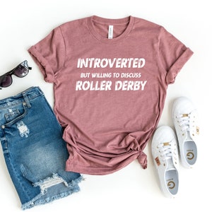 May include: A mauve t-shirt with the text "Introverted but willing to discuss roller derby" in white lettering.