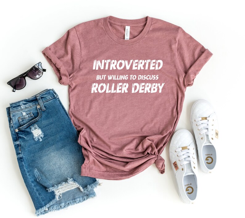 May include: A mauve t-shirt with the text "Introverted but willing to discuss roller derby" in white lettering.