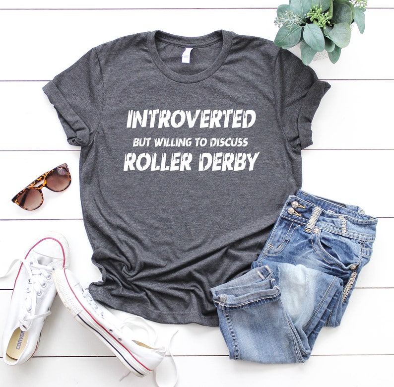 May include: A dark gray t-shirt with white text that reads "Introverted but willing to discuss roller derby". The shirt is laid flat on a white wooden surface with a pair of jeans, sunglasses, and white sneakers.