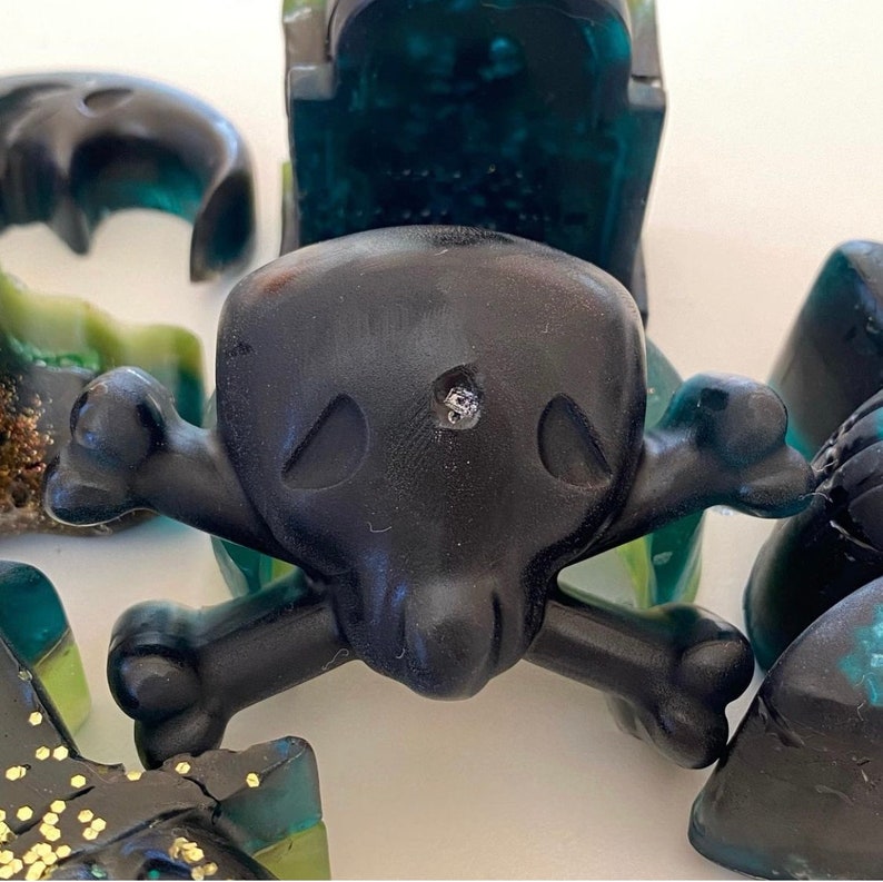Puede incluir: Una figura de calavera y huesos cruzados de color negro y verde con un tachuela plateada en el centro del cr&aacute;neo. La figura est&aacute; hecha de un material liso y brillante.