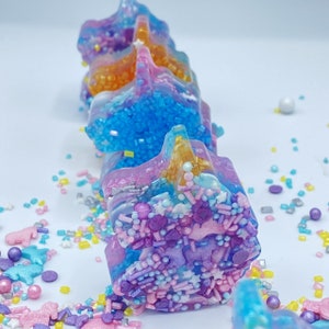 May include: A row of colorful, star-shaped resin decorations. The decorations are layered with shades of blue, pink, and gold, and are covered in sprinkles and small decorative shapes. The background is white.
