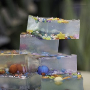 Puede incluir: Una pila de cuatro bloques de resina transparentes, cuadrados y transl&uacute;cidos, con chispas de colores y otros elementos decorativos incrustados. Los bloques est&aacute;n apilados sobre una superficie beige claro.