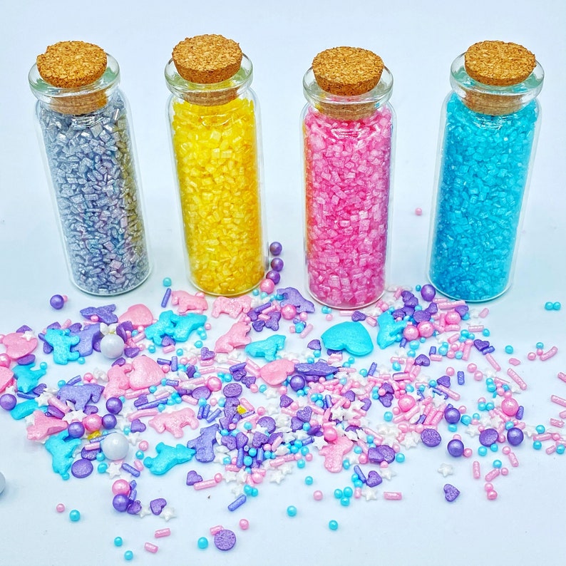 May include: Four small glass bottles with cork stoppers, filled with colorful sprinkles. The sprinkles are in shades of silver, yellow, pink, and blue. Scattered around the bottles are various sprinkles in the shapes of hearts, stars, and animals, in pastel colors.