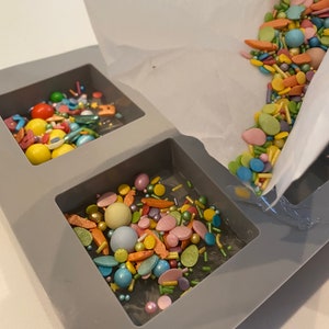 Puede incluir: Un molde de silicona gris con dos compartimentos llenos de chispas y caramelos de colores. Las chispas se est&aacute;n vertiendo desde una bolsa de papel blanca.