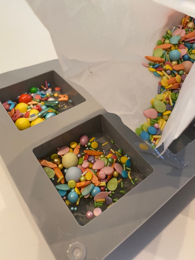 Puede incluir: Un molde de silicona gris con dos compartimentos llenos de chispas y caramelos de colores. Las chispas se est&aacute;n vertiendo desde una bolsa de papel blanca.