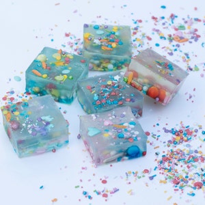 Puede incluir: Seis postres de gelatina cuadrados de color azul claro con chispas y cuentas de colores en su interior. Los postres est&aacute;n sobre una superficie blanca con chispas esparcidas a su alrededor.