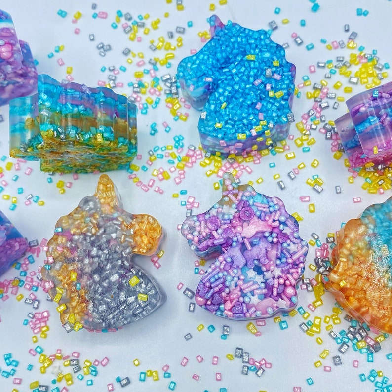 May include: A collection of colorful, unicorn-shaped resin art pieces. The pieces are decorated with sprinkles and glitter in shades of blue, purple, gold, and silver. The background is white with scattered sprinkles.