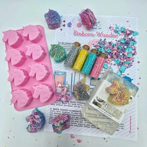 May include: A pink silicone unicorn mold, several glitter-covered unicorn-shaped soaps, and small glass bottles filled with glitter. The image also includes a sheet of paper with German text and a scattering of sprinkles.