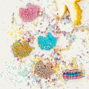 May include: An overhead shot of colorful, glitter-covered resin shapes, including a bow, a bird, and a rectangle. The shapes are scattered on a white surface, surrounded by sprinkles in pastel colors and a decorative unicorn.