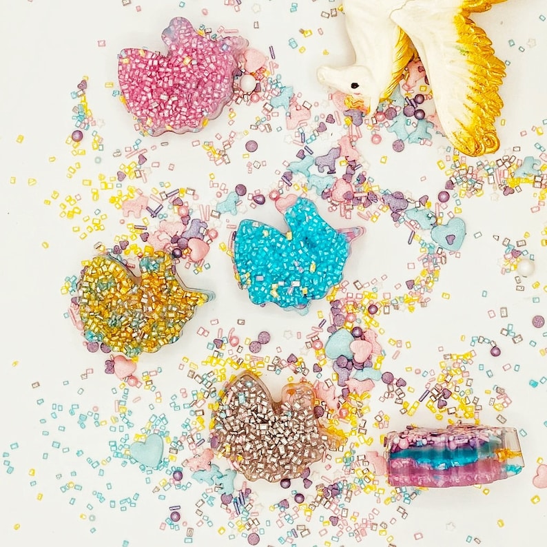 May include: An overhead shot of colorful, glitter-covered resin shapes, including a bow, a bird, and a rectangle. The shapes are scattered on a white surface, surrounded by sprinkles in pastel colors and a decorative unicorn.