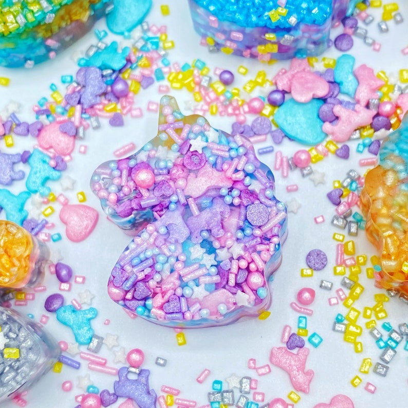 May include: A close-up of a unicorn-shaped confection, decorated with colorful sprinkles and edible glitter. The unicorn is a mix of pastel colors, including pink, purple, and blue. Other unicorn-shaped treats are visible in the background.