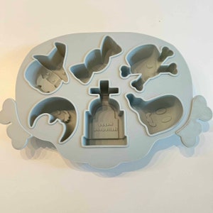 Puede incluir: Bandeja de cubitos de hielo de Halloween de silicona gris con moldes para un fantasma, un murci&eacute;lago, un bast&oacute;n de caramelo, una calavera y huesos cruzados, una l&aacute;pida con el texto "Happy Halloween" y un sombrero de bruja.
