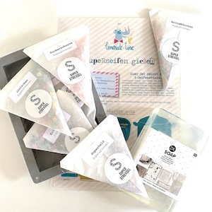 Puede incluir: Un juego de ocho jabones hechos a mano con una variedad de colores y texturas. Los jabones est&aacute;n empaquetados en bolsas de papel blanco con un logotipo en blanco y negro que dice "Super Streusel". Los jabones est&aacute;n dispuestos sobre una superficie blanca con un fondo a rayas blanco y amarillo. El texto "Euer Set reicht f&uuml;r 8 Seifenst&uuml;cke" es visible en el fondo.