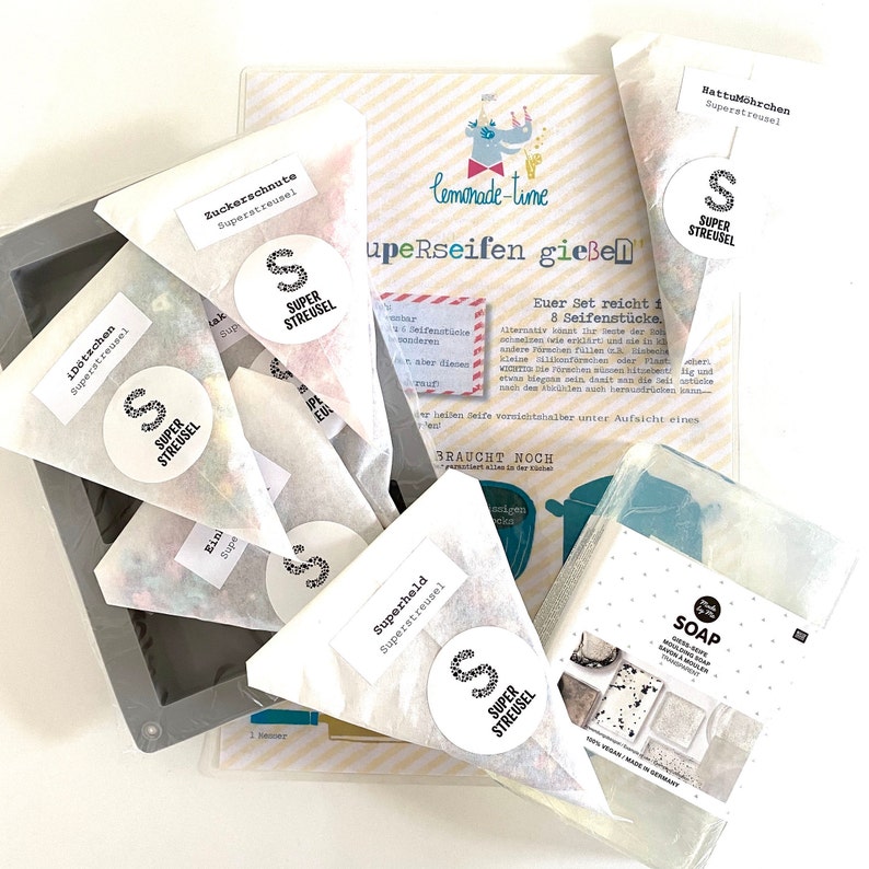 Puede incluir: Un juego de ocho jabones hechos a mano con una variedad de colores y texturas. Los jabones est&aacute;n empaquetados en bolsas de papel blanco con un logotipo en blanco y negro que dice "Super Streusel". Los jabones est&aacute;n dispuestos sobre una superficie blanca con un fondo a rayas blanco y amarillo. El texto "Euer Set reicht f&uuml;r 8 Seifenst&uuml;cke" es visible en el fondo.