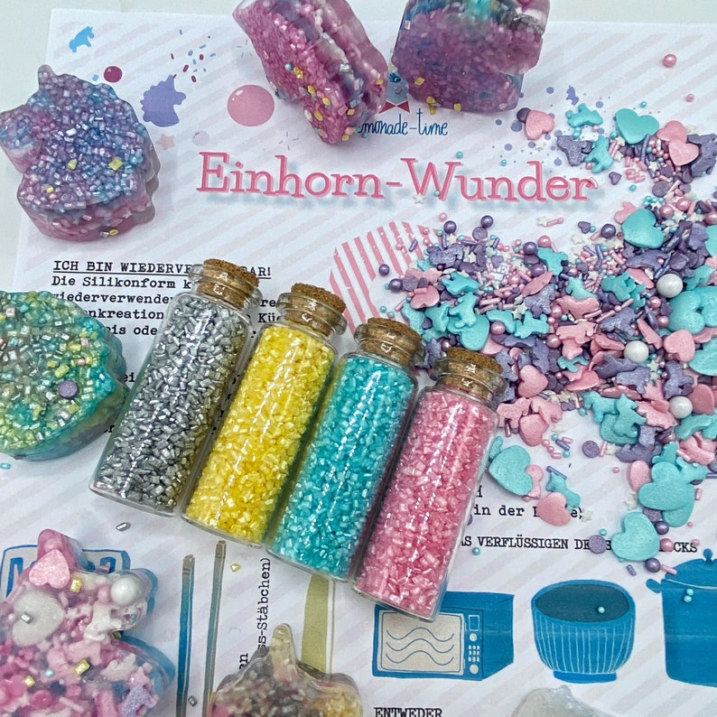 May include: A collection of craft supplies with a unicorn theme. The image features small glass bottles filled with colorful sprinkles, unicorn-shaped molds, and a variety of sprinkles in pastel colors. The text "Einhorn-Wunder" is visible.