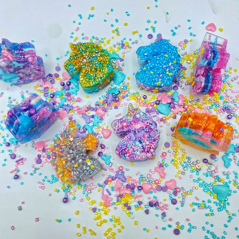 May include: An assortment of colorful unicorn-shaped decorations. Each unicorn is a different color, including blue, pink, orange, and green, and is covered in sprinkles and other decorative elements. The decorations are scattered on a white surface.
