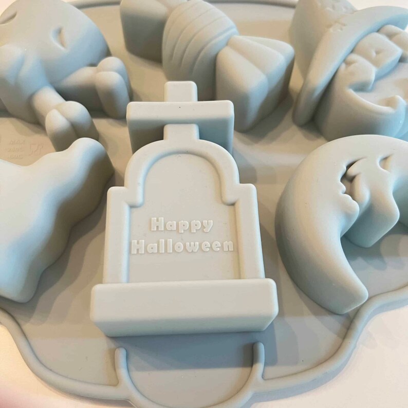Puede incluir: Un molde de silicona azul claro con seis formas diferentes con tem&aacute;tica de Halloween: un fantasma, un sombrero de bruja, un bast&oacute;n de caramelo, una l&aacute;pida con el texto "Happy Halloween", una luna creciente con una cara de fantasma y un esqueleto con huesos cruzados.