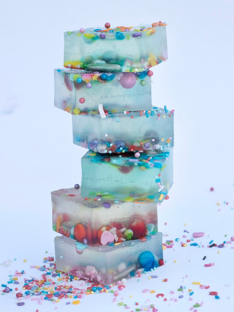 Puede incluir: Una pila de cinco caramelos transparentes, cuadrados y gelatinosos con chispas y caramelos coloridos en su interior. Los caramelos est&aacute;n apilados sobre una superficie blanca con chispas esparcidas a su alrededor.