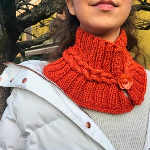 Könnte beinhalten: Ein orangefarbener Strick-Halsschmeichler mit Zopfmuster und Blumenakzent. Der Schal ist aus dickem, strukturiertem Garn gefertigt und soll den Hals warm halten. Ein gemütliches Winteraccessoire.