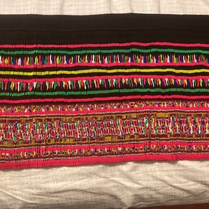 May include: A close-up of a colorful fabric with intricate embroidery. The fabric features a black background with red, green, yellow, and blue threads woven into a geometric pattern.