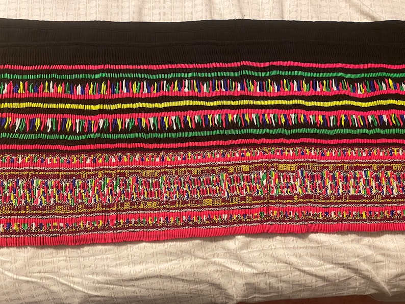 May include: A close-up of a colorful fabric with intricate embroidery. The fabric features a black background with red, green, yellow, and blue threads woven into a geometric pattern.
