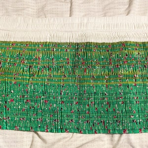 May include: A green fabric with a white and green floral pattern is pleated and gathered at the top. The fabric is folded in half and laid flat on a white surface.