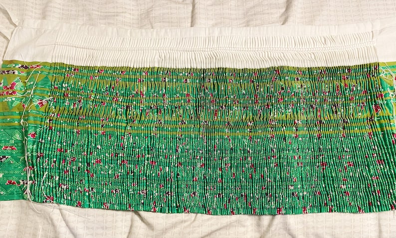 May include: A green fabric with a white and green floral pattern is pleated and gathered at the top. The fabric is folded in half and laid flat on a white surface.