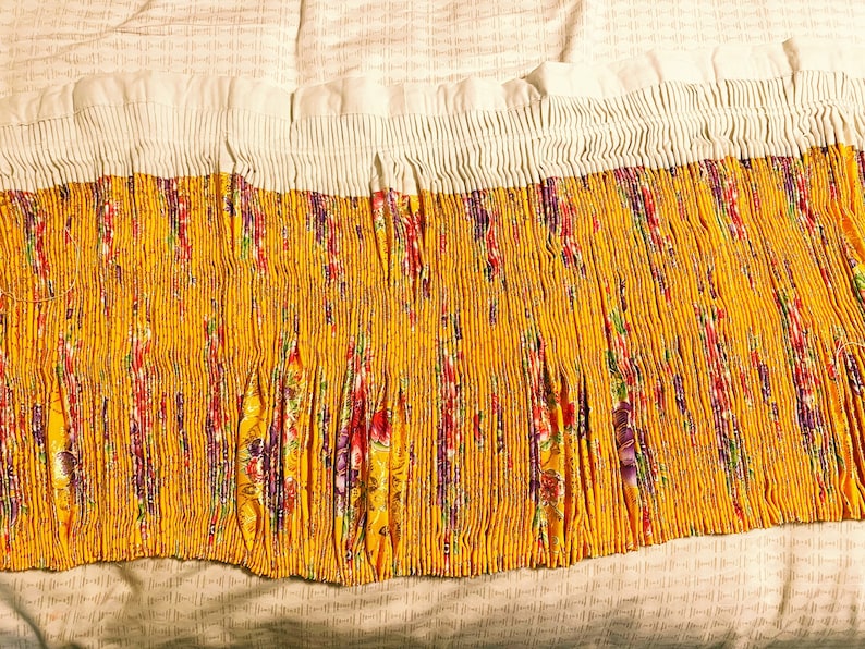 May include: A yellow fabric with a floral pattern is pleated and gathered at the top. The fabric is white at the top and has a ruffled edge.