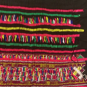 May include: A close-up of a colorful fabric with intricate embroidery. The fabric is black with a pattern of pink, green, yellow, blue, and white threads. The embroidery is in a geometric pattern and is very detailed.