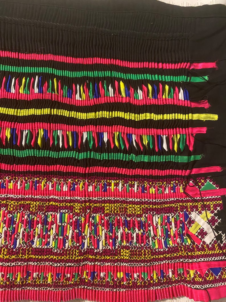 May include: A close-up of a colorful fabric with intricate embroidery. The fabric is black with a pattern of pink, green, yellow, blue, and white threads. The embroidery is in a geometric pattern and is very detailed.