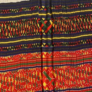 May include: A close-up of a colorful fabric with a repeating geometric pattern. The fabric is made up of red, yellow, green, blue, and black threads. The pattern is symmetrical and intricate.