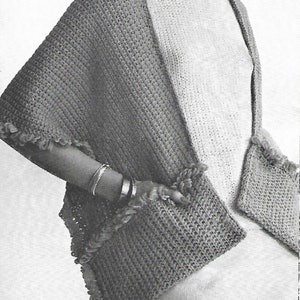 Pode incluir: Um xale de crochê cinza com detalhes de franjas e dois bolsos. O xale é usado por uma modelo com cabelo curto e escuro.