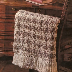 Peut inclure: Une couverture en crochet blanche et marron avec une bordure en franges. La couverture est drapée sur le dossier d'une chaise en bois.