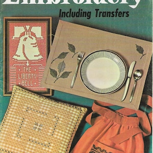 May include: A vintage embroidery book cover featuring a green background with a red, white and blue Liberty Bell design, a beige placemat with embroidered leaves, a white plate with a black rim, a beige pillow with a brown cross-stitch design, and an orange apron with a brown embroidered design. The book title is "Embroidery Including Transfers" and the book number is 129. The price is 29 pence.