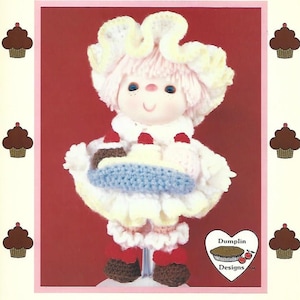 May include: A crochet doll dressed as a banana split. The doll has a white and yellow hat, pink hair, and a blue and white dress. The doll is holding a crocheted banana split. The doll is standing on a stand. The image includes the text "Banana Split" and "Dumplin Designs".