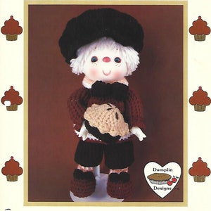 May include: Crochet pattern for a pecan pie doll. The doll is wearing a black hat, a brown sweater, brown pants, and brown shoes. The doll is holding a pie in its hands. The pattern is from "Dumplin Designs".