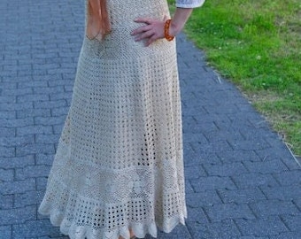 May include: A woman wearing a white blouse with a brown scarf and a long, crocheted beige skirt. She is wearing a brown belt and a brown bracelet. She is standing on a brick path.