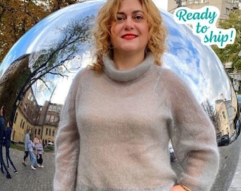 Può includere: Una donna indossa un maglione color grigio chiaro, semitrasparente, a collo alto. Il maglione ha una trama a coste e maniche lunghe. La donna è in piedi davanti a una grande sfera argentata riflettente. Il testo "Ready to ship!" è scritto in blu turchese su sfondo bianco.