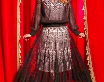 May include: A two-piece outfit consisting of a long-sleeved black knit top with a sheer, intricate openwork pattern and a matching midi-length skirt featuring a black lace overlay with a scalloped hem over a solid light-colored lining.
