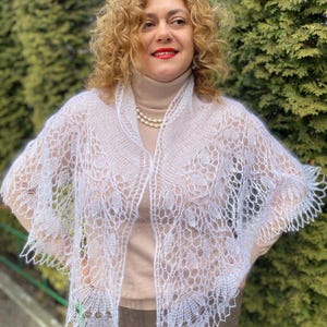 Puede incluir: Un chal blanco de tejido abierto con un delicado estampado floral. El chal está drapeado sobre los hombros, mostrando su intrincado diseño y textura aireada. Los bordes del chal están festoneados, añadiendo un toque decorativo a la prenda.