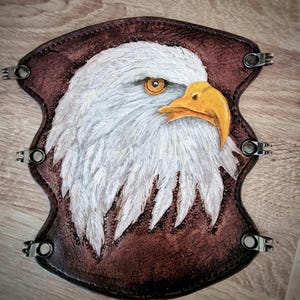 Könnte beinhalten: Ein braunes Lederstück mit einem darin geschnitzten weißen Kopf eines Weißkopfseeadlers. Das Auge des Adlers ist gelb und sein Schnabel ist orange. Das Stück hat Metallnieten an den Rändern.