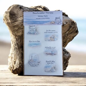Pode incluir: Um pacote de autocolantes "Beachcomber Therapy" com seis autocolantes de vinil. Cada autocolante apresenta ilustrações com temas de praia e frases como "The beach is my therapy" e "Find Calm by the Sea". O pacote é exibido em madeira flutuante.