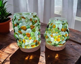 May include: Two glass jar candle holders, decorated with colorful sea glass in shades of green, yellow, and brown. The jars sit on wooden bases, and the light shines through the sea glass, creating a warm glow. The jars are on a wooden table.