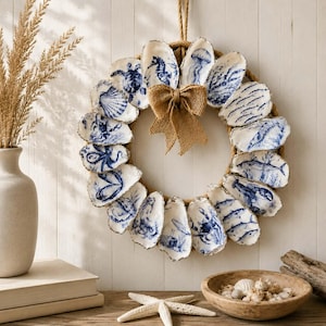 Puede incluir: Una corona circular hecha de cuerda natural, adornada con numerosas conchas de ostra blancas que presentan intrincadas ilustraciones azules de vida marina, incluyendo un caballito de mar, medusa y cangrejo, con un lazo central de arpillera.