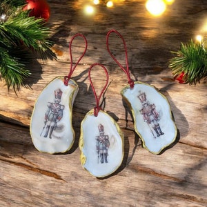 May include: Three Christmas ornaments made from oyster shells with gold trim. Each ornament features a vintage illustration of a toy soldier in red and blue.