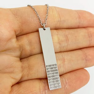 Puede incluir: Un collar con colgante de barra plateada y cadena fina. El colgante rectangular tiene código binario grabado. El collar se muestra en la mano de una persona.